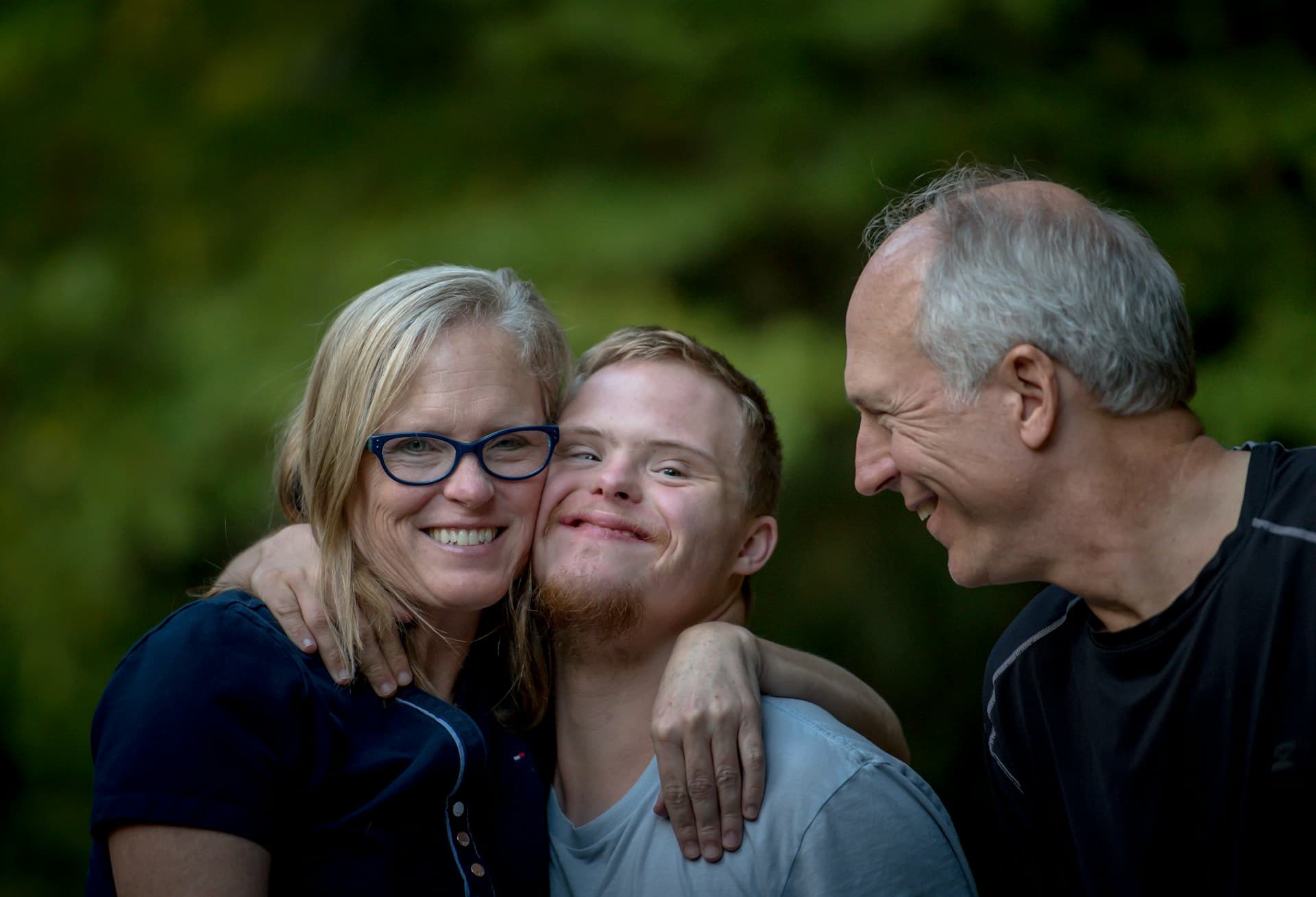 A person with disability is being hugged by a woman while a man smiles at them.