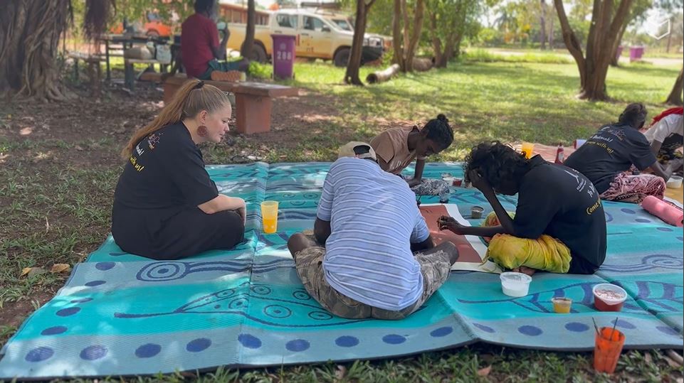 A behaviour support practitioner sits on a blue mat with First Nations artists painting outside, engaging with a participant’s community and support network. In the background is tree trunks, dappled sunlight and grass